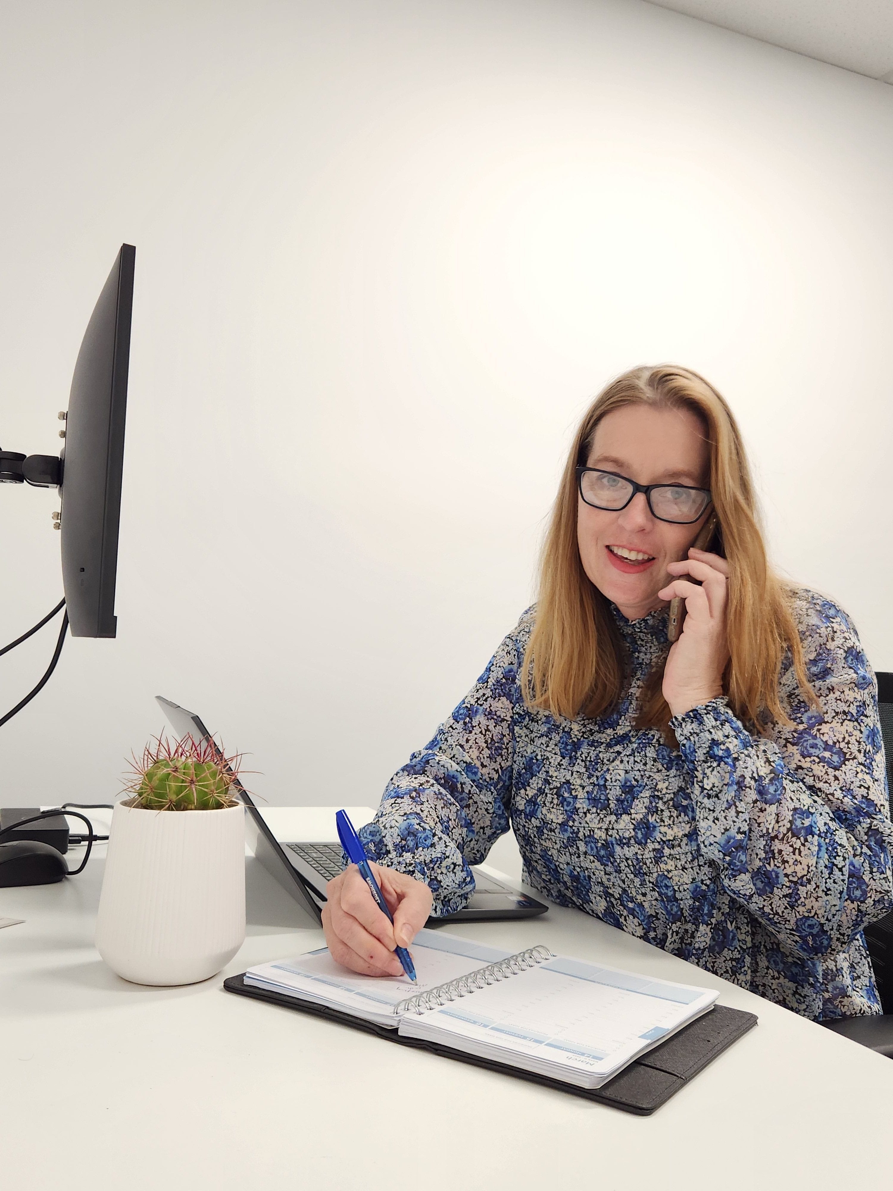 Helen sitting a a desk writing in her diary and talking on the phone