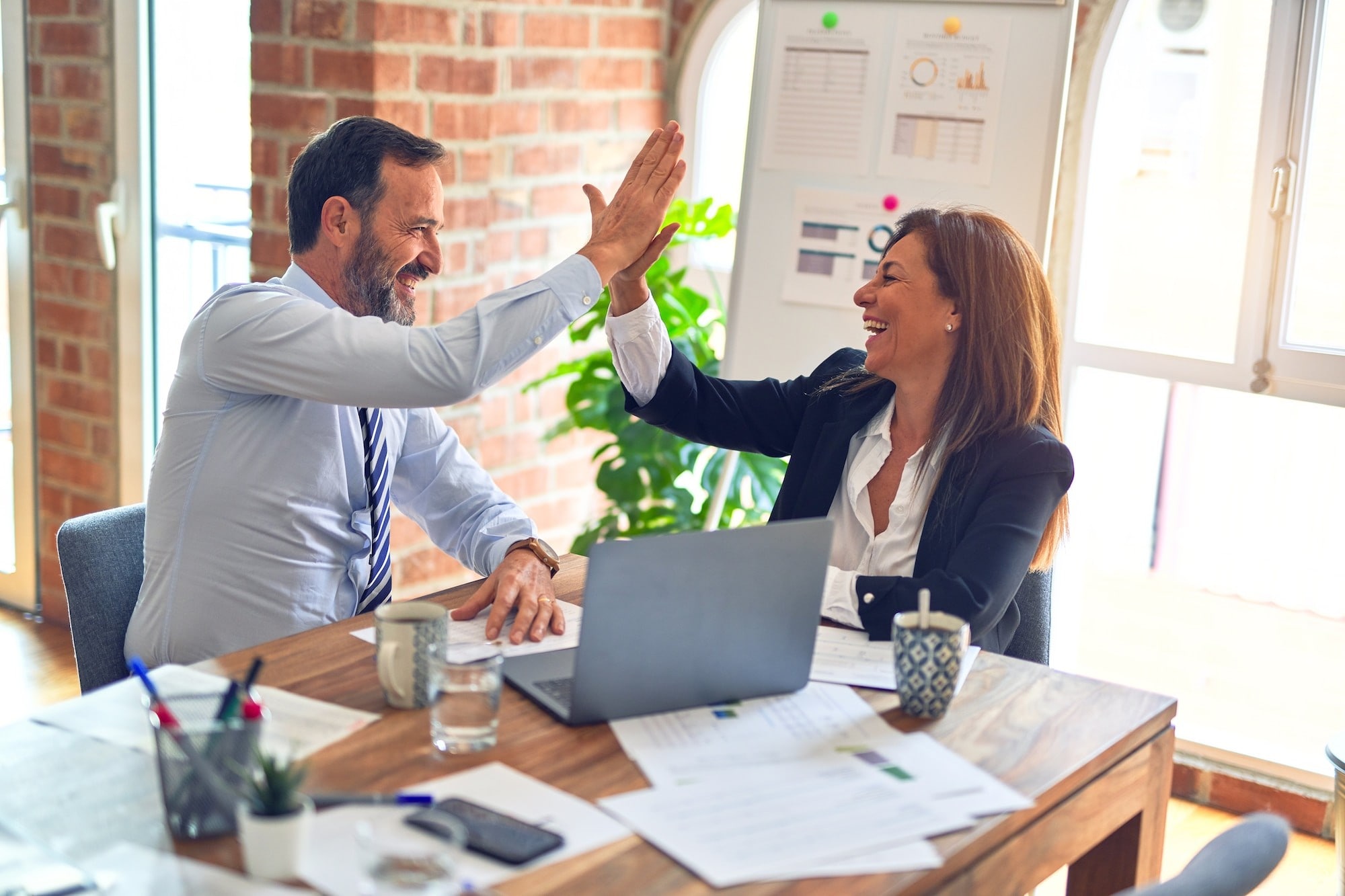 A male and female sitting a desk in front of a computer with paperwork and coffees on the desk doing a high five